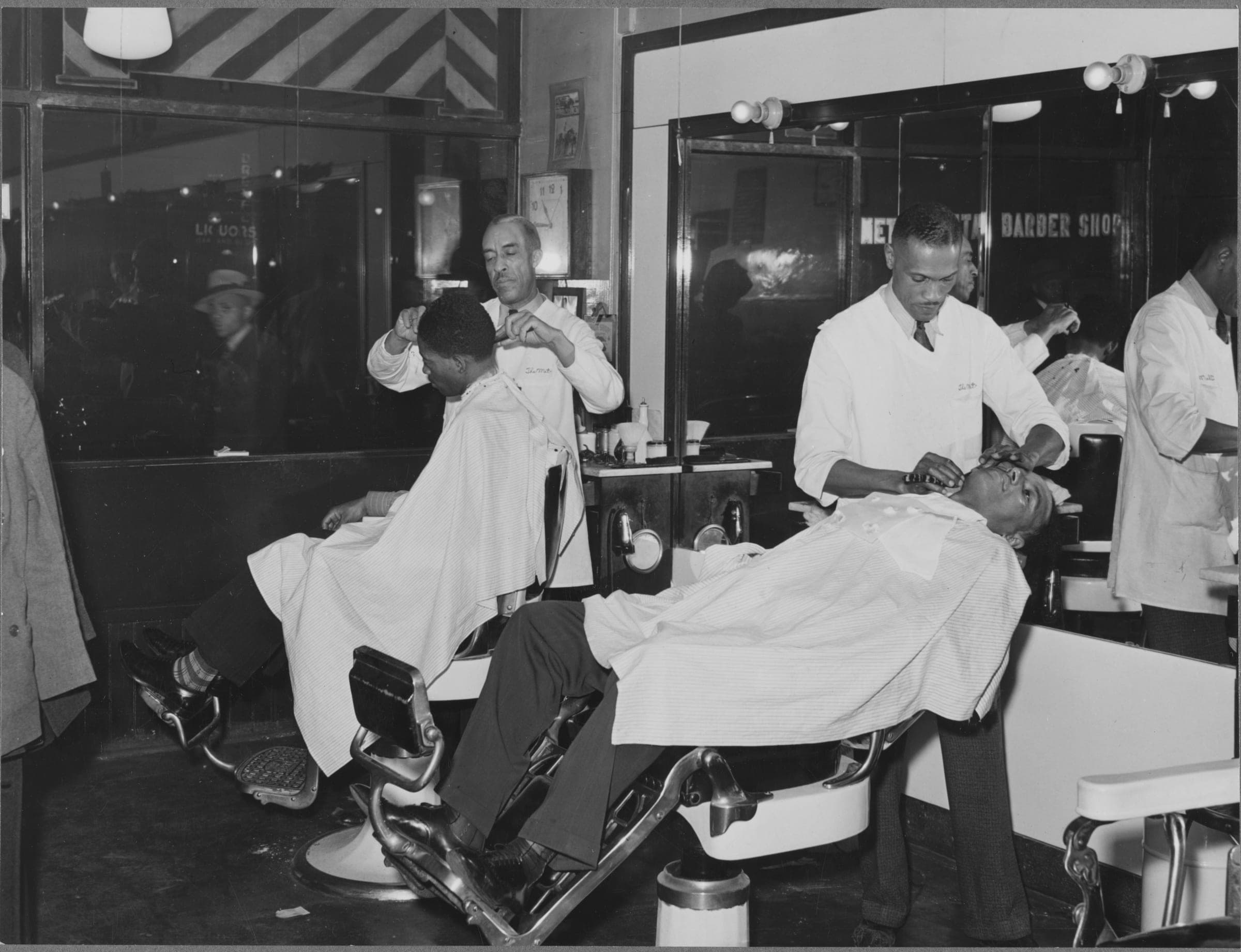 Inside a barbershop at night: two barbers in white coats work on two customers in reclined chairs, the shop's name lettered across the mirror behind them and the street showing through the window on the left.