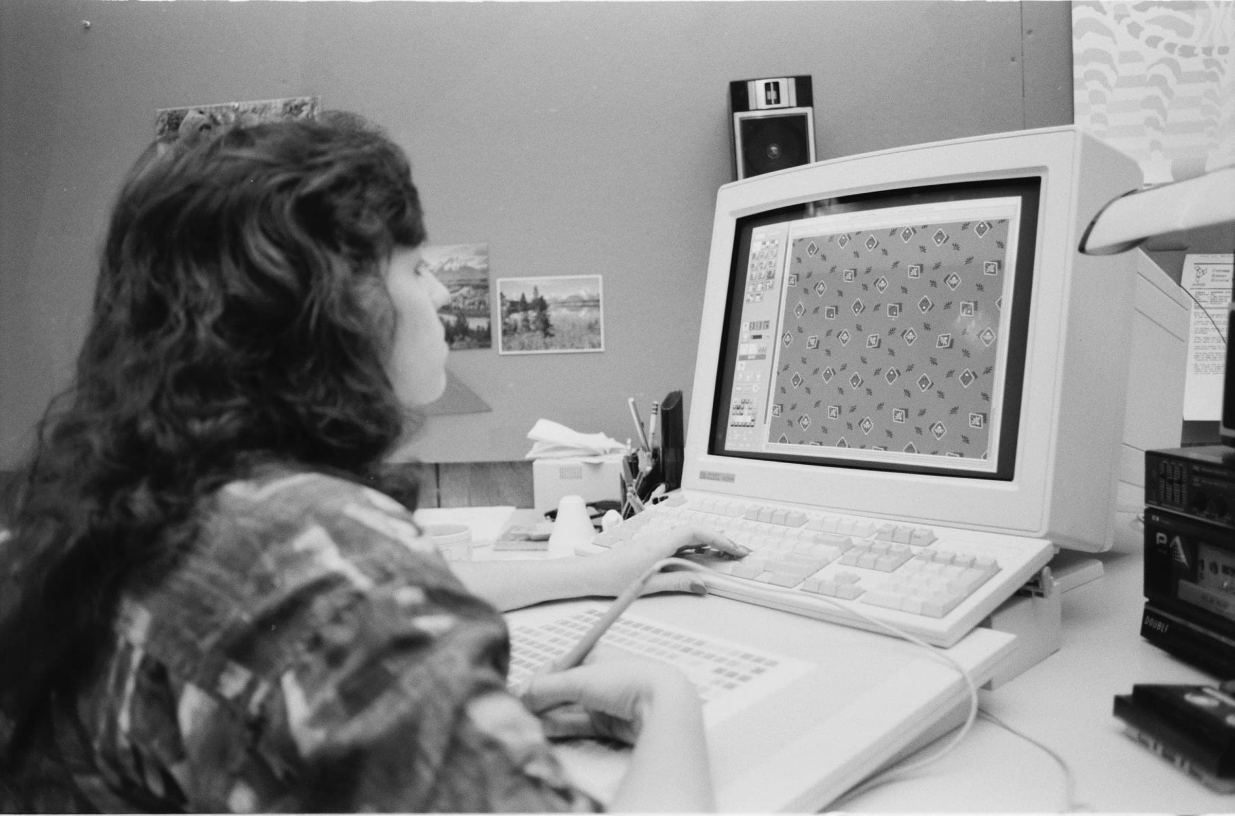 A fabric designer seen from behind at her desk, long dark hair over a patterned blouse, her hands on a keyboard and a graphics tablet, a repeating diamond pattern filling the monitor in front of her.