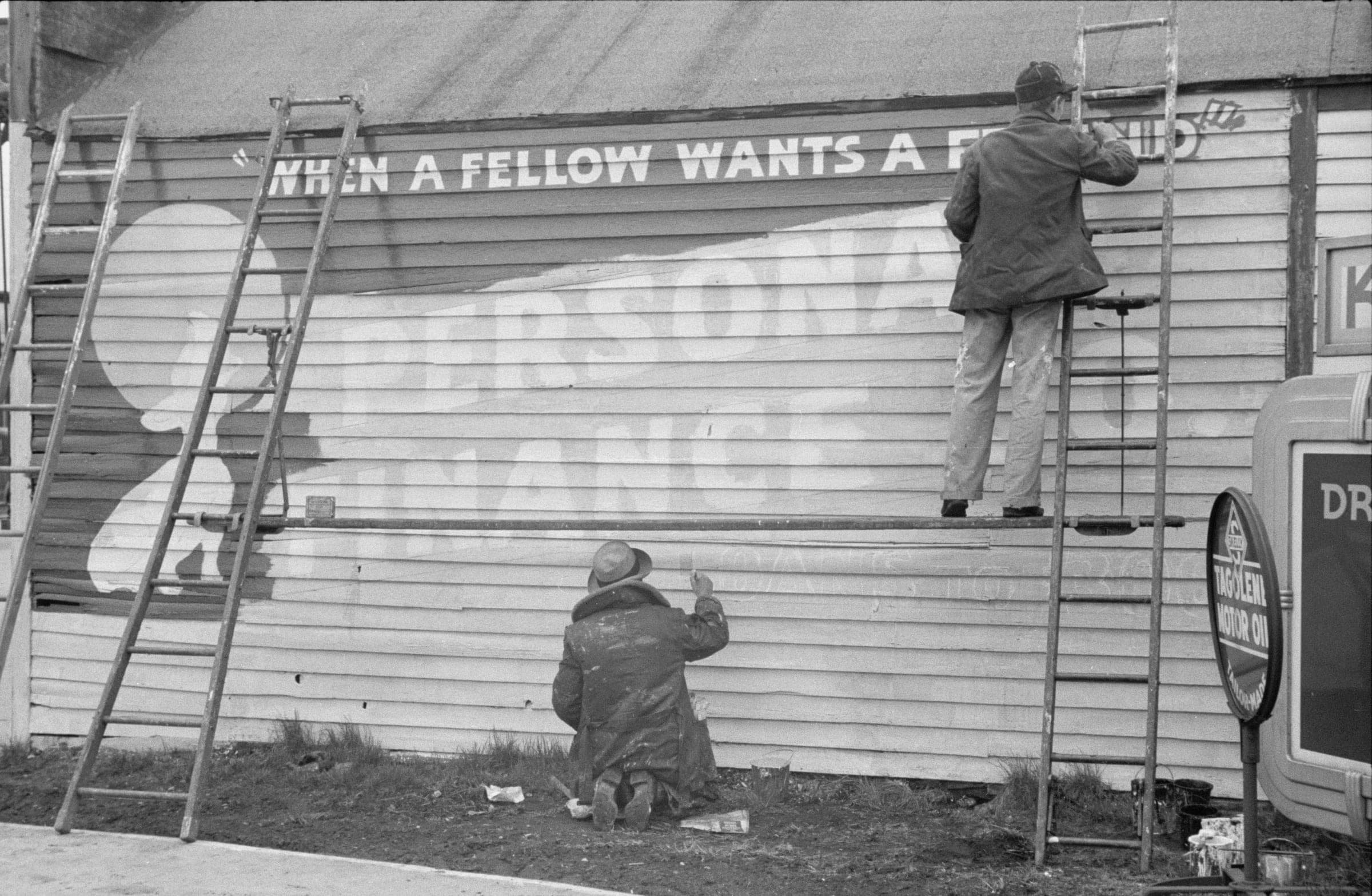 Two sign painters at work on the clapboard side of a building: one kneeling on the ground lettering the lower line, one up a ladder on the right, between them the half-painted words PERSONAL FINANCE and a line above reading WHEN A FELLOW WANTS A FRIEND.